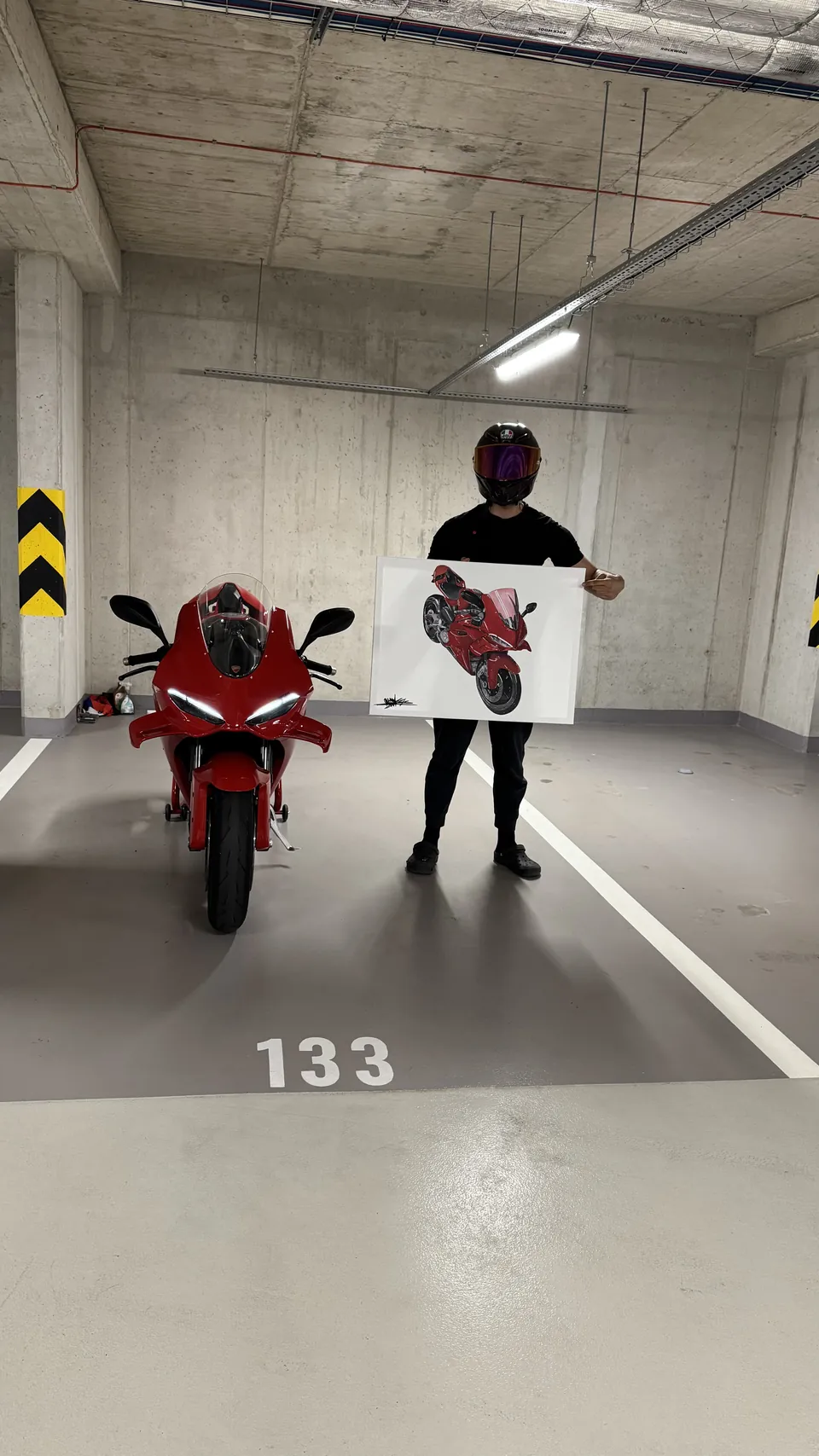 Jezdec v helmě drží obraz červené motorky Ducati vedle skutečného stroje v garáži